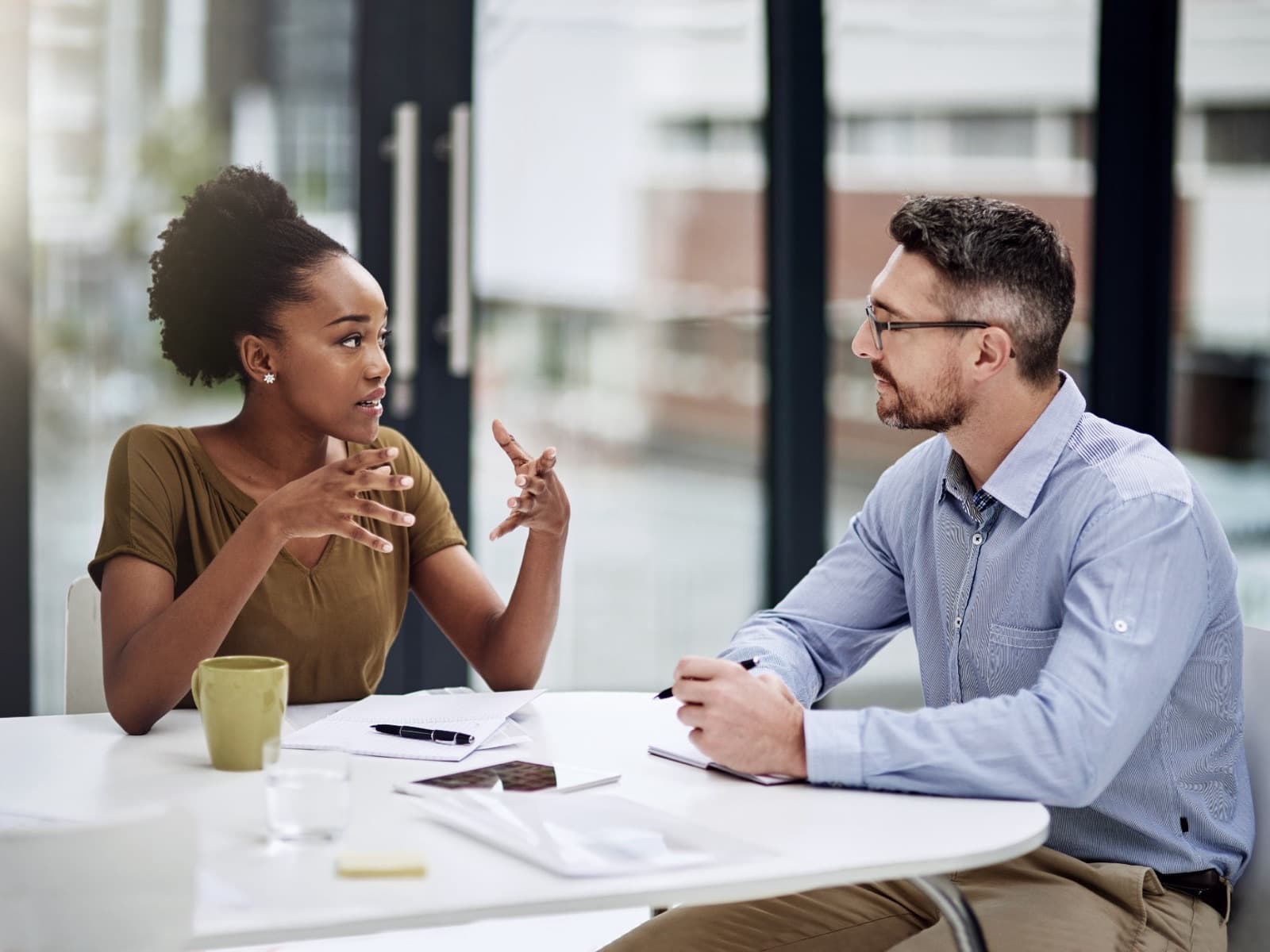 Two colleagues in conversation across a table, one gesturing mid-sentence while the other reads their hands and expression